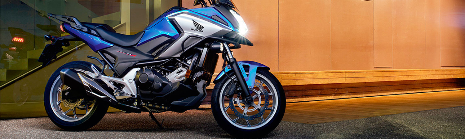 A stylish blue and silver Honda Motorcycle parked outside Ridenow Chandler, its headlights cutting through the dusk, looking sleek.